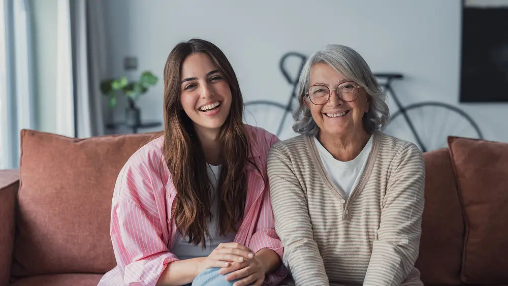 A young woman in a pink striped shirt sits smiling next to an older woman with gray hair and glasses, both on a brown couch in a brightly lit living room.