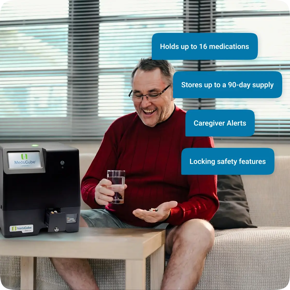 A man in a red sweater sits on a couch beside a MedaCube medication dispenser, holding pills and a glass.