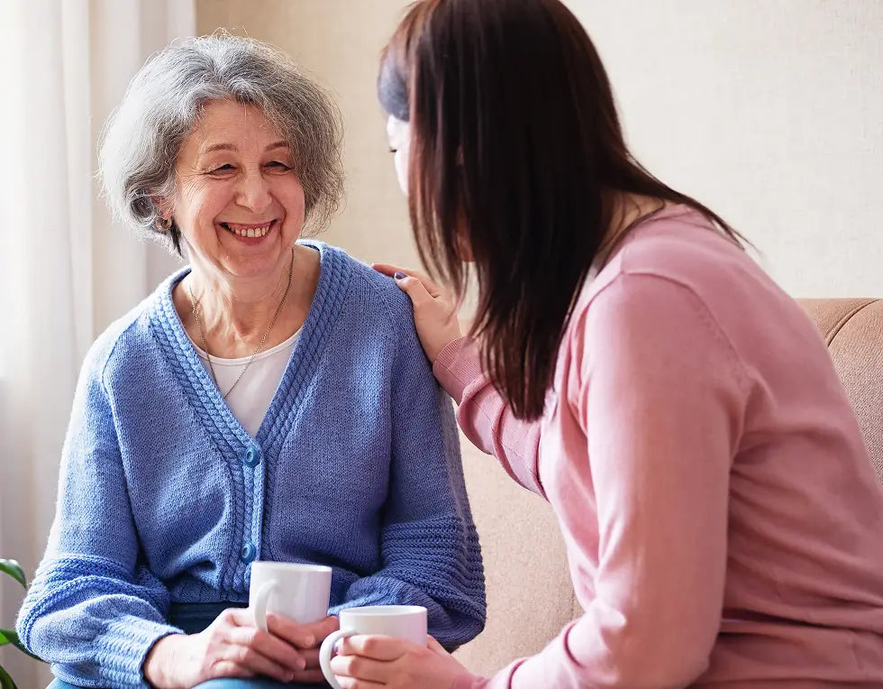 An elderly woman with gray hair smiles warmly while holding a white mug, seated next to a younger woman with dark hair in a pink top who is leaning in to speak. They are on a beige couch in a brightly lit indoor setting.